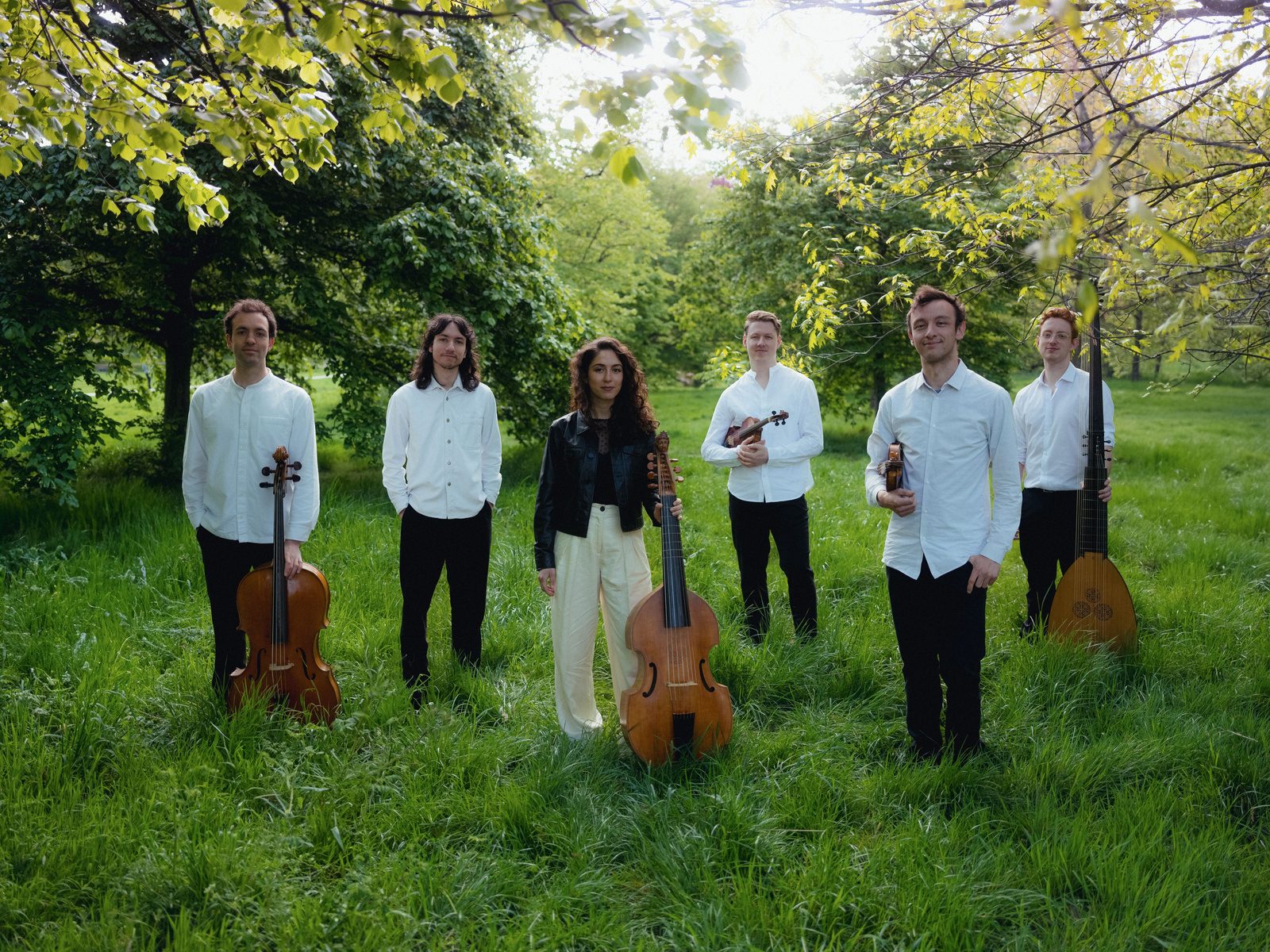 The six members of Bellot Ensemble standing outdoors among trees, holding period instruments including viols and a theorbo.
